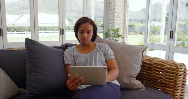 Woman sitting on comfortable sofa using a tablet in a bright, modern living room with large windows. Ideal for themes related to technology, modern lifestyle, remote work, leisure activities, home comfort, relaxation, and indoor living. Suitable for blogs, advertisements, and articles focusing on home tech use, digital lifestyle, and home decor.