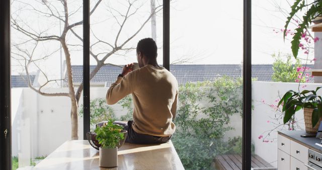 Man sitting on a table by a large window looking outside in a modern home. Ideal for emphasizing relaxation, peaceful living, morning routines or lifestyle blog content about home decor, relaxation, and personal space.