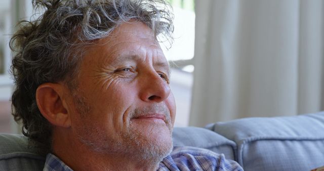 Older man with gray curly hair sitting on a sofa, looking thoughtful and relaxed. Can be used in campaigns about retirement, mental well-being, and relaxation or for illustrating mature lifestyle concepts.