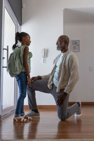 African grandfather kneeling and smiling while greeting granddaughter with backpack at door