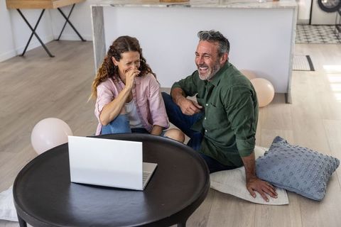 Middle-aged couple laughing during virtual celebration at home with laptop and balloons