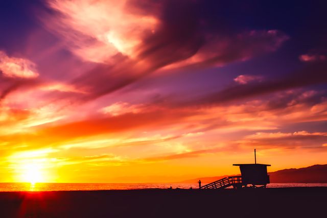 This breathtaking scene showcases an awe-inspiring sunset at the beach with a lifeguard tower silhouetted against the colorful sky. Ideal for use in travel brochures, beach resort advertisements, relaxation or mindfulness content, summer promotion materials, sunset-themed posters, and any project requiring a serene, vibrant, and coastal aesthetic.