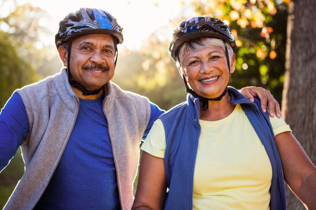 Senior couple smiling while enjoying a cycling trip in a park. They are wearing helmets and casual clothing, indicating a healthy and active lifestyle. This image can be used for promoting senior health, fitness programs, outdoor activities, and family bonding.