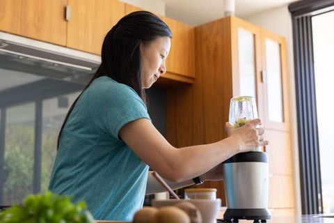 Mature asian woman blending green smoothie in sunlit modern kitchen