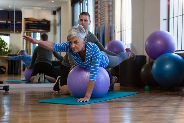Senior woman performing exercise on fitness ball with assistance from physiotherapist in clinic. Ideal for illustrating physical therapy, rehabilitation, senior fitness, and health and wellness programs. Useful for medical and fitness websites, brochures, and articles on elderly care.