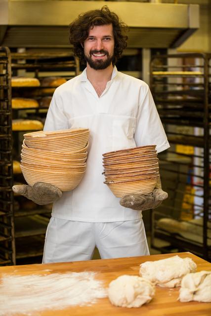 Smiling baker holding a stack of bowls while preparing dough in a bakery. Ideal for use in articles or advertisements related to baking, culinary arts, professional chefs, and the food industry. Can also be used for promoting bakery products, cooking classes, or culinary schools.
