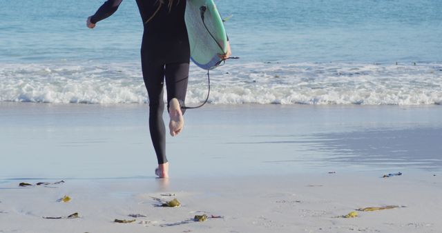 A person runs towards the ocean holding a surfboard while wearing a wetsuit. This captures a sense of excitement and anticipation often associated with water sports. Ideal for use in advertising for surfing equipment, beach-related activities, and fitness.