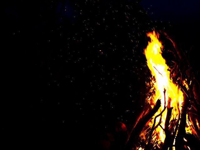 Intense bonfire blazing against a pitch-black night sky with glowing embers and visible sparks. Ideal for illustrating concepts of outdoor activities, camping, warmth, survival skills, and night-time gatherings. Perfect for use in travel blogs, camping guides, survival tutorials, and nature-themed projects.