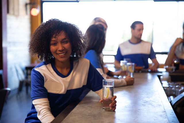 Portrait of biracial woman at the bar in a pub during the day, wearing team shirt holding a drink her friends in the background. Friendship leisure time fun.