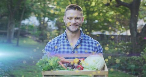 Smiling man holding wooden crate of fresh organic vegetables in backyard garden dappled sunlight