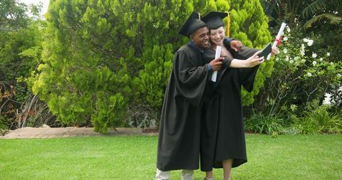 Graduates posing for selfie on campus lawn holding diplomas in caps and gowns, smiling