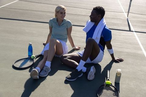 Diverse tennis partners resting on hard court after match, smiling and hydrating