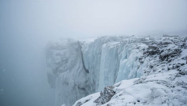 Towering frozen icefall at snow-covered cliff edge, foggy glacial precipice rugged rocks