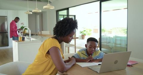 African american mother helping son with homework at bright kitchen table using laptop
