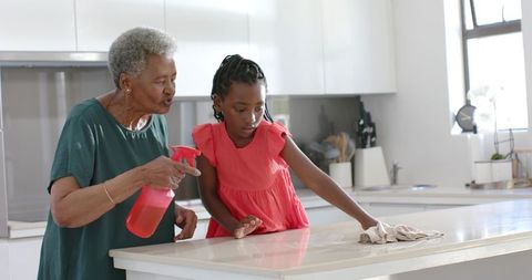 Elderly woman teaching young girl cleaning kitchen countertop in bright modern home