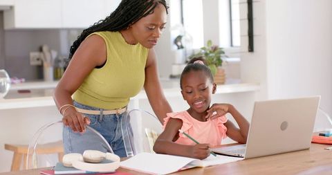 African american mother guiding daughter with homework at kitchen table using laptop