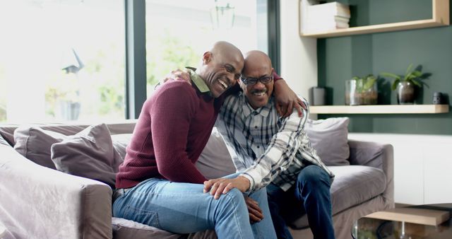 Father and son are sitting on a comfortable couch in a contemporary living room. They are smiling, cuddling, and enjoying their time together, showing a strong family bond. This image can be used to depict family life, love, and togetherness in a modern home setting. Ideal for advertisements and content focusing on family values, therapy and counseling, and home decor.