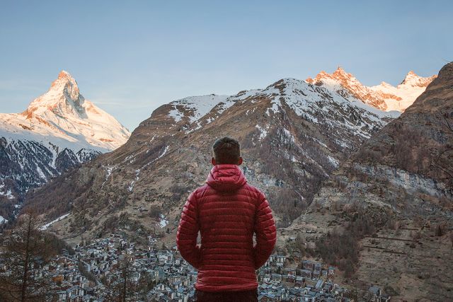 Man in warm jacket enjoying the serene beauty of Swiss mountains during sunrise, with snowy peaks and a quaint village below. Perfect for topics like adventure, outdoor activities, travel destinations, and connecting with nature.