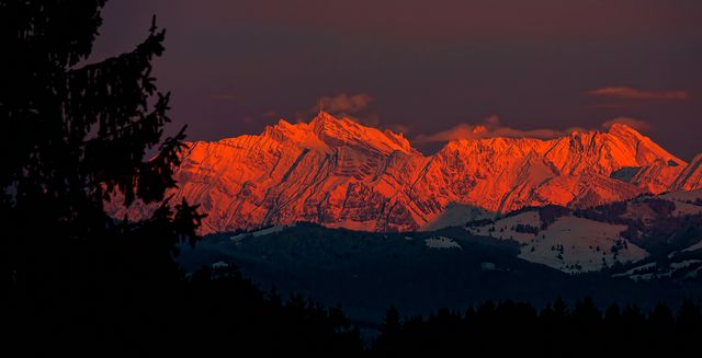 Snow-covered mountain peaks bathed in warm red light during sunset with dark forest silhouettes in foreground. Ideal for use in nature photography, travel blog, postcards, and winter-themed designs.