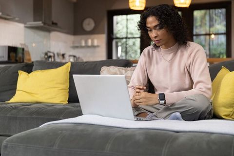 Asian non-binary person working remotely on laptop in cozy modern living room with kitchen view