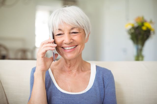 Senior woman with short gray hair talking on mobile phone, smiling warmly. She is sitting on a couch in a bright living room with a bouquet of flowers in the background. Ideal for use in advertisements or articles about senior lifestyle, communication technology for the elderly, or home living.