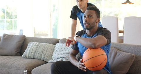 Diverse friends watching basketball on sofa holding ball wearing smartwatch casual home
