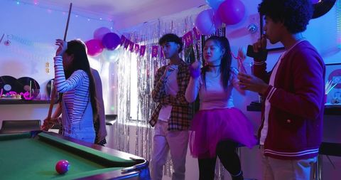 Diverse friends playing pool at colorful birthday party with balloons and tinsel curtain