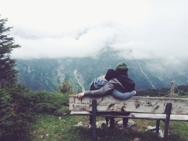 Couple sitting on wooden bench embracing and enjoying foggy mountain scenery. This peaceful and romantic image can be used for travel blogs, nature-related advertisements, couple's retreats promotions, and adventure-themed marketing materials.