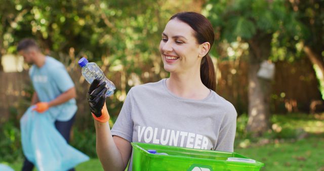 Smiling caucasian woman wearing volunteer t shirt holding recycling crate, collecting plastic waste. eco conservation volunteers doing countryside clean-up.