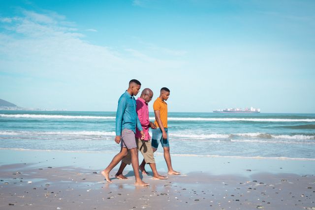 African american senior father and twin adult sons walking and talking at beach. healthy outdoor family leisure time together.