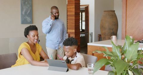 African american family helping child using tablet at kitchen table while father on phone