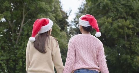 Female friends wearing santa hats holding hands outdoors in cozy sweaters for holiday season