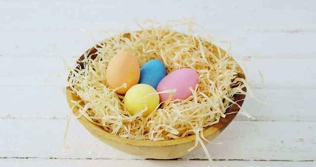A wooden bowl contains colorful Easter eggs nestled in straw, with copy space. These pastel-colored eggs symbolize the festive spirit of the Easter holiday.
