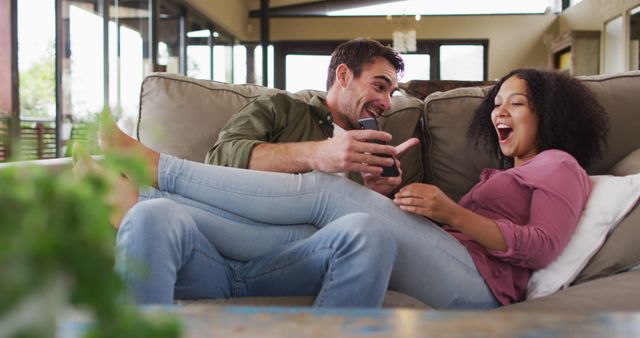Biracial couple smiling while looking in smartphone together on the couch at vacation home. couple honeymoon and vacation concept