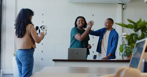 African American coworkers high-fiving at bright coworking counter, laptop headphones team