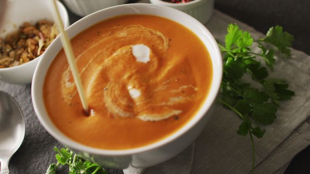 Cream pouring into a bowl of tomato soup, creating a swirling effect. Fresh cilantro on the side adds a touch of green. This video is perfect for illustrating food blogs, recipe websites, and culinary promotions highlighting comforting meals and seasonal dishes.