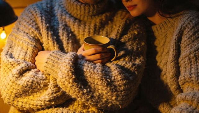 Couple cuddling in chunky knit sweaters holding ceramic mug under warm lamp glow