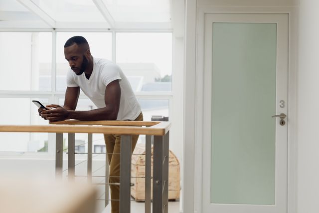 This image depicts an African-American man focused on a smartphone while standing inside a bright modern interior with white walls and wooden railings. Suitable for themes related to technology, communication, mobile devices, or modern lifestyles. Ideal for promotional materials, websites, blogs, and ads targeting a tech-savvy audience or demonstrating a contemporary environment.