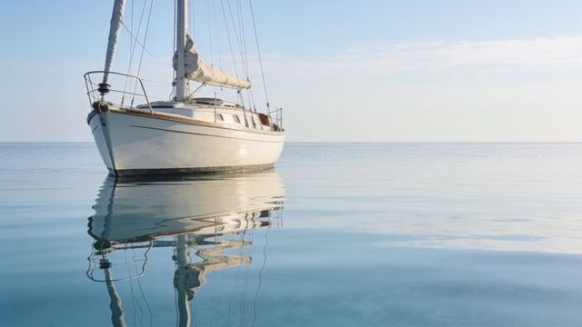 Sailboat floating on calm sea with mirror reflection and soft morning sky horizon
