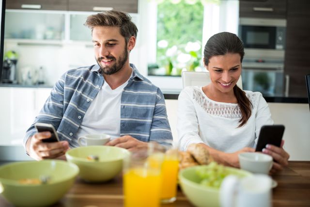 Couple sitting at dining table in kitchen, both using mobile phones while having breakfast. Bowls of food and glasses of orange juice on table. Ideal for illustrating modern lifestyle, technology use, and daily routines. Suitable for articles on digital communication, family life, and breakfast habits.
