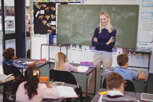Teacher standing in front of classroom, helping students with their homework. Blackboard in background with alphabet letters. Ideal for educational content, school-related articles, teaching resources, and academic websites.