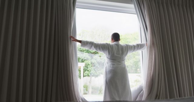 Individual in bathrobe opening curtains in hotel room, looking out at the serene greenery. Ideal for blogs on travel, relaxation, luxury living, hospitality advertisements, and hotel promotions showcasing comfortable settings and peaceful environments.