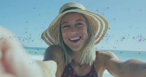 Smiling young woman taking beach selfie wearing straw hat and maroon swimsuit by turquoise sea