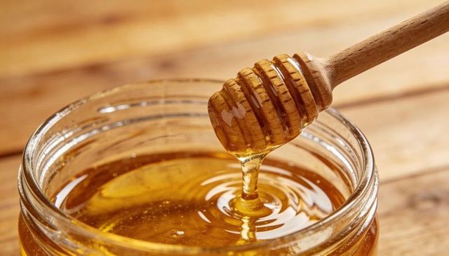 Golden Honey Dripping from Wooden Dipper into Glass Jar with Ripples and Warm Glow