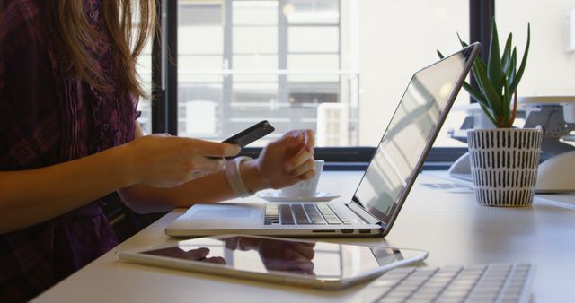 Talented businesswoman managing multiple tasks in a modern office environment, holding smartphone and coffee cup while typing on laptop. This image is perfect for depicting productivity, technology use in professional settings, workplace dynamics, or details about modern office life.