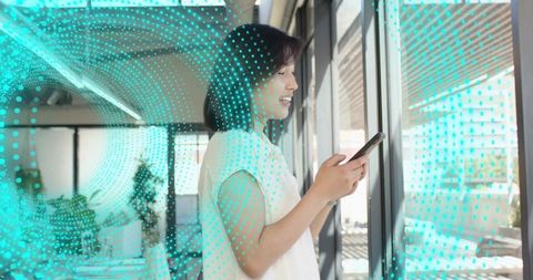 Smiling woman checking smartphone by office window with teal digital data overlay
