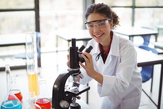 Young schoolgirl wearing safety goggles and lab coat, smiling while using a microscope in a science laboratory. Various colorful chemical solutions and laboratory glassware are on the table. Ideal for educational content, STEM promotion, science classroom materials, and academic publications.