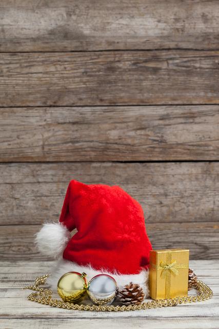 This festive scene features a Santa hat, Christmas ornaments, a small gift box, pinecones, and a golden chain atop a rustic wooden table. Perfect for holiday-themed advertisements, greeting cards, seasonal blog posts, or social media content showcasing the Christmas spirit.