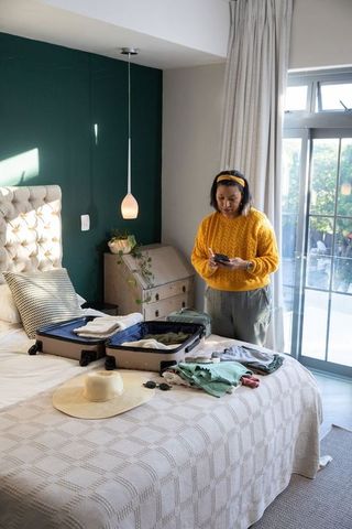 Mature woman packing suitcase in sunlit bedroom while using smartphone for travel planning