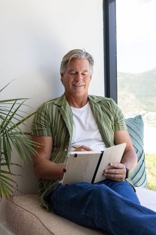 Senior man writing in notebook on sunlit window bench, relaxed home journaling
